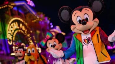 Mickey Mouse, sporting disney pride merch—a rainbow-colored jacket with a heart—stands before Minnie Mouse, Goofy, and Pluto at a vibrant, illuminated theme park parade at night.