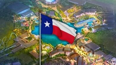 A large Texas state flag waves in the foreground, with an illuminated amusement park featuring rides, buildings, and water attractions visible in the background at Universal Kids Resort.