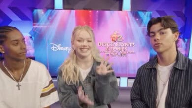 Three young people sit together in front of a colorful Disney “Descendants Zombies Worlds Collide Tour” backdrop. The girl in the middle is smiling and waving, while the two boys on either side look towards the camera.