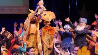 Children in colorful costumes perform a scene from "The Lion King" musical on stage, showcasing the excitement of Disney Musicals in Schools as one child holds up a stuffed lion and others gesture joyfully around them.