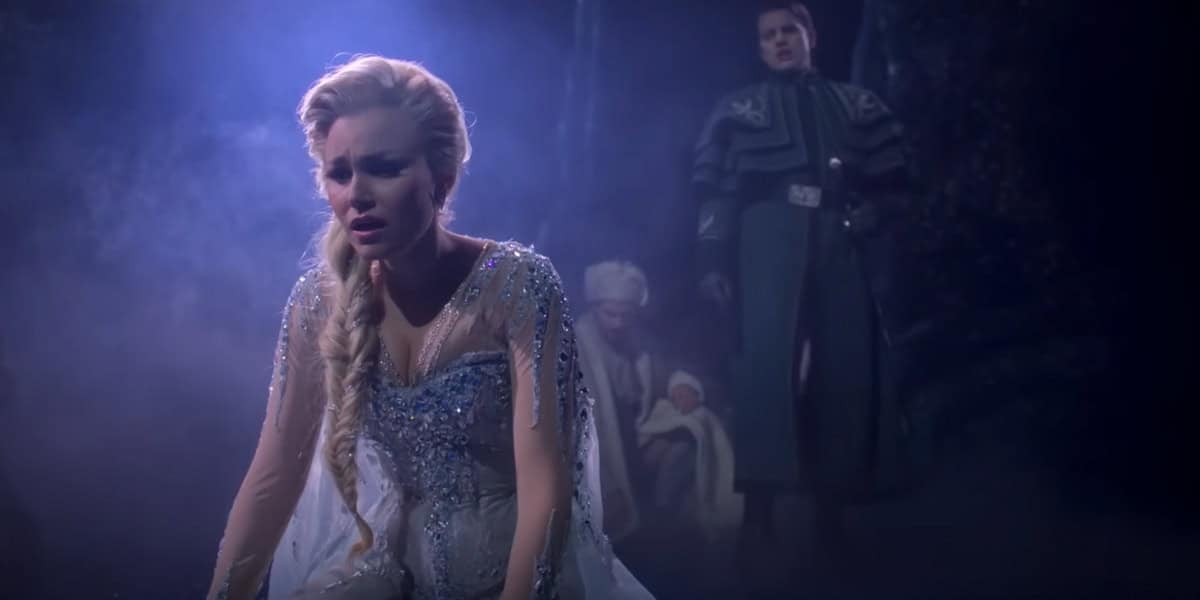 A woman in a sparkling, ice-blue dress kneels on the ground, looking distressed. In the misty background, two people stand, one in dark clothing and another holding a baby. The scene is dimly lit and dramatic.