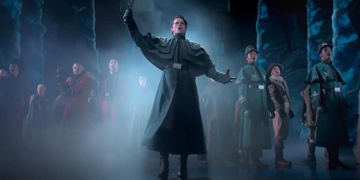 A group of actors in dramatic costumes stand on stage amid blue-lit fog; the central figure in a dark green robe with an arm raised, while others in uniform stand in formation behind. The setting appears theatrical and intense.
