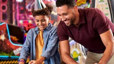 A smiling child wearing a party hat and a man play an arcade game together at Chuck E Cheese. Bright lights and arcade machines fill the background, creating a festive and playful atmosphere.