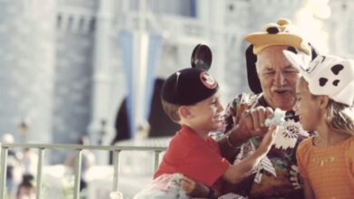 An elderly man and two children wearing Disney character hats laugh and share a treat in front of a castle at a theme park. The scene is joyful and playful.
