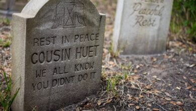 A gravestone reads “Rest in Peace Cousin Huet. We all know you didn’t do it,” in a grassy cemetery, with another faded gravestone visible in the background.