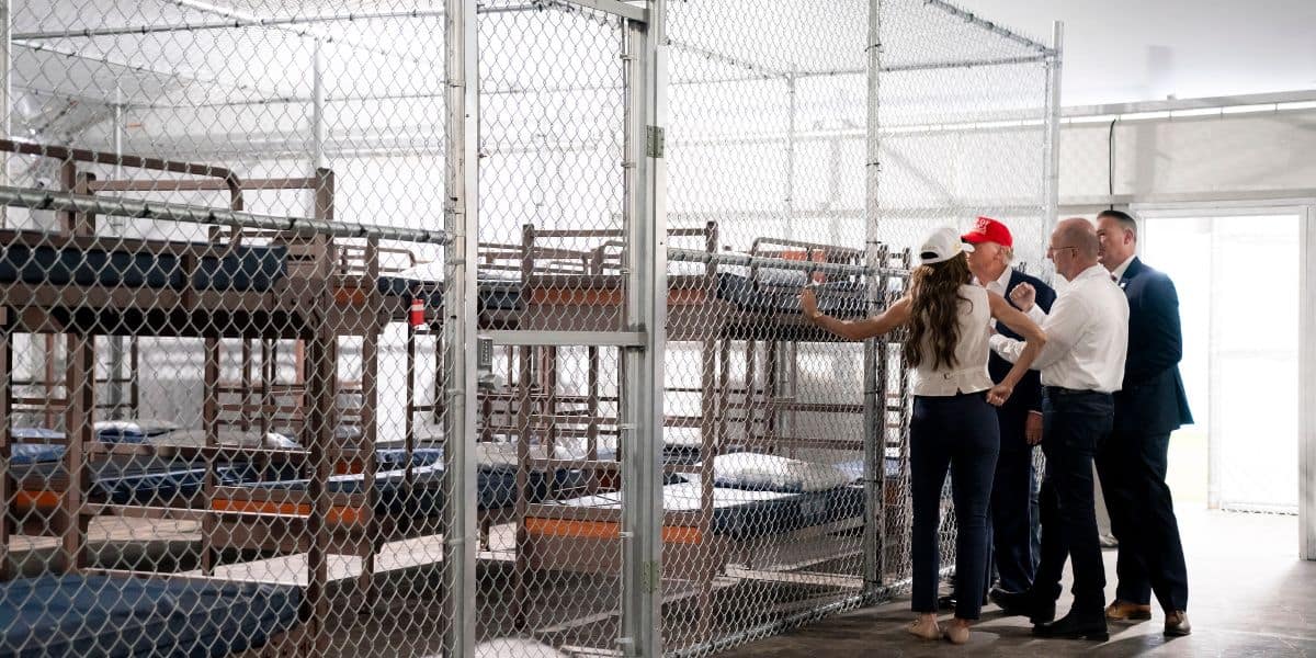 Four people stand inside a fenced enclosure with metal bunk beds and thin mattresses, appearing to inspect or discuss the facility. The space is stark, with minimal bedding and chain-link fencing.