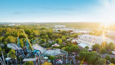 A sunny aerial view of an amusement park with roller coasters, green trees, a large white dome building, and clear blue skies. The sun is low on the horizon, casting a warm glow over the scene at Hersheypark.