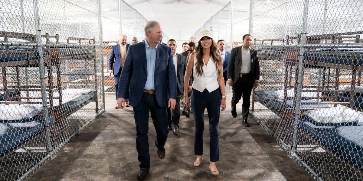 A group of people, including a man in a blue blazer and a woman in a white top and hat, walk between rows of bunk beds inside a facility with chain-link fencing and concrete floors.