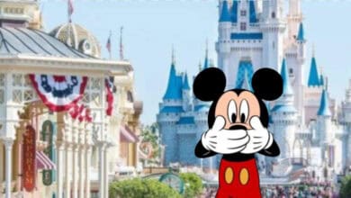Mickey Mouse covers his mouth with both hands in front of Main Street and Cinderella Castle at a Disney theme park, decorated with patriotic banners.
