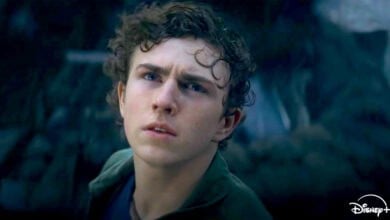 A young man with curly hair and a serious expression looks upward in a dim, dramatic outdoor setting. He appears wet, drops of water on his face hinting at adventure from the Percy Jackson Season 2 trailer. The Disney+ logo is visible in the bottom right corner.