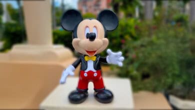 A figurine of Mickey Mouse with a yellow bow tie, red shorts, and white gloves is standing outside with greenery and buildings blurred in the background.