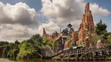 A roller coaster train on a wooden track winds through rugged, rocky terrain and greenery near a lake under a partly cloudy sky.