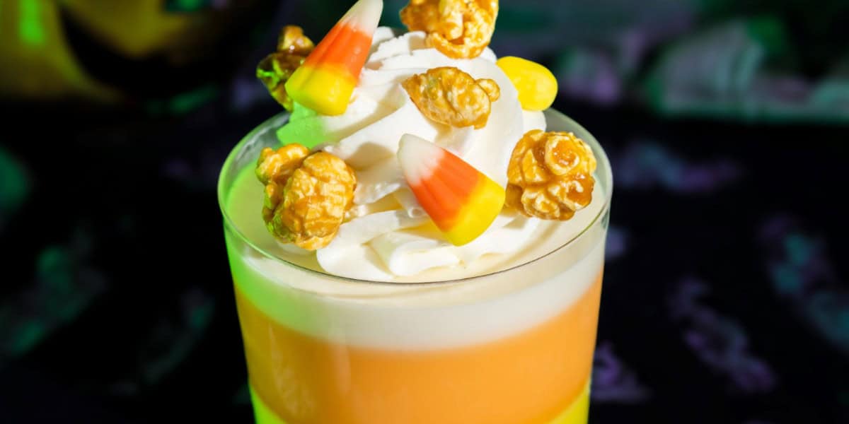 A magical, Disney-inspired layered dessert in a glass with yellow, orange, and white sections, topped with whipped cream, candy corn pieces, and caramel popcorn.