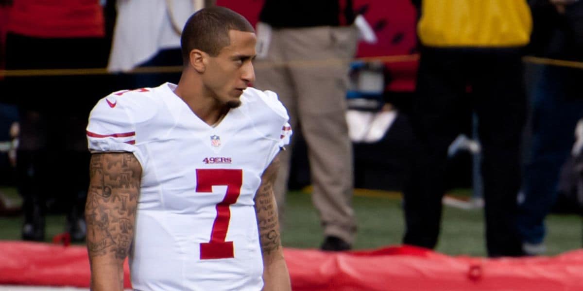 A football player wearing a white San Francisco 49ers uniform with the number 7 stands on the field, looking to the side. There are people and a red sideline banner in the background.