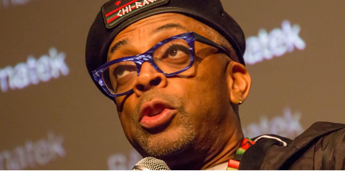 A man wearing blue glasses, a black beret with "CHI-RAQ" written on it, and a colorful necklace speaks into a microphone against a blurred background with indistinct text.