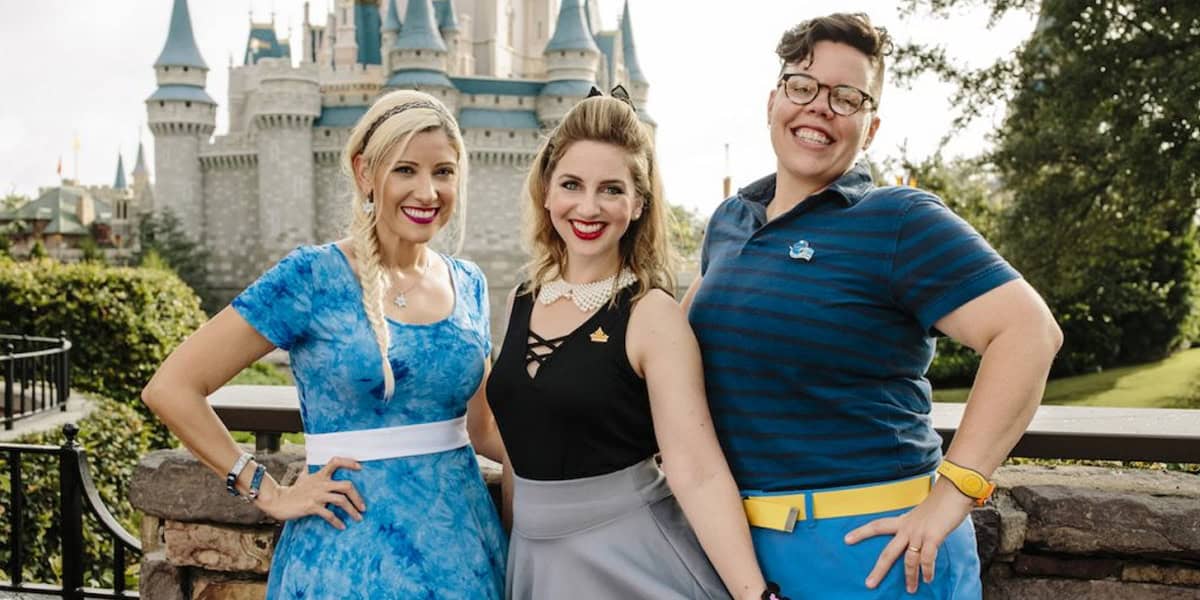 Three smiling adults pose in front of a fairytale castle at a theme park, wearing colorful outfits and accessories, with greenery and blue sky in the background.