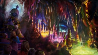 People ride a mine train through a colorful, glowing cave filled with stalactites, stalagmites, and pools of vibrant light, creating a magical, fantastical atmosphere inside the cavern.