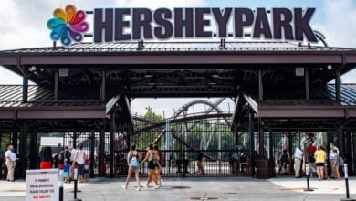 Entrance to Hersheypark with people walking in and out; a large Hersheypark sign is above the gate, and a roller coaster is visible in the background at this theme park news.