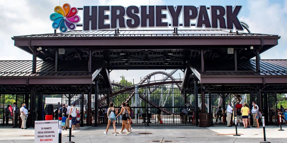 Entrance to Hersheypark with people walking in and out; a large Hersheypark sign is above the gate, and a roller coaster is visible in the background at this theme park news.
