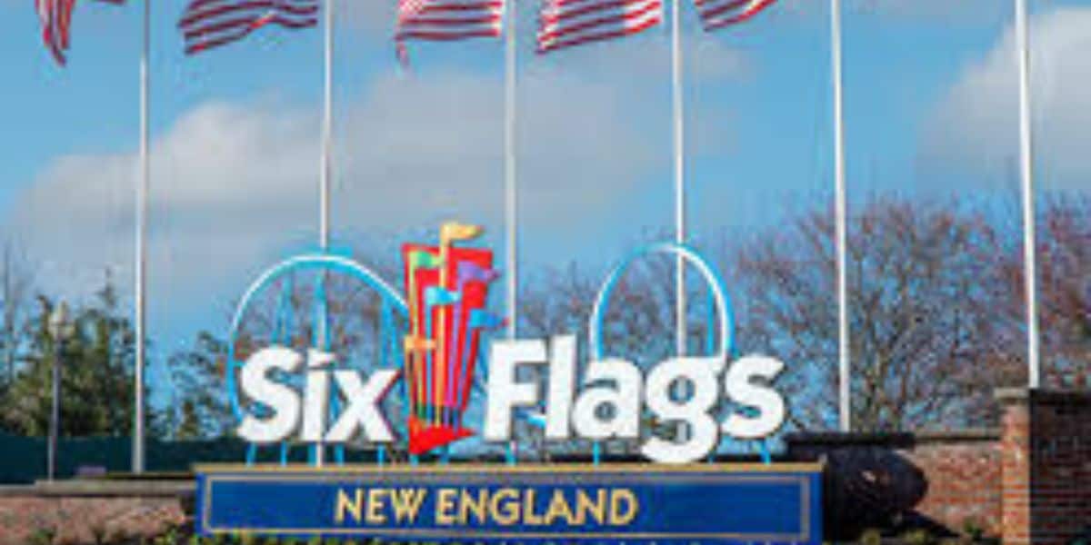 A sign for Six Flags New England stands in front of several waving American flags, with trees and a blue sky in the background.
