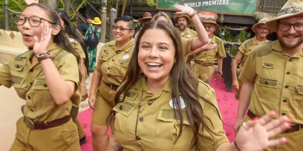 A group of people in matching khaki uniforms smile and wave as they walk on a pink carpet outdoors, with greenery and a "World Premiere" sign in the background.