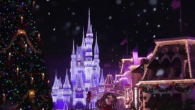 A brightly lit christmas castle stands in the background. Snow falls gently over a festive street with garlands, wreaths, and a large, colorful Christmas tree on the left.
