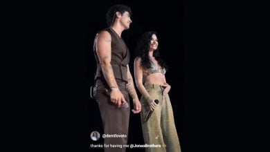 Two singers stand on stage in front of a black background, smiling and holding microphones. One wears a sleeveless brown outfit, and the other wears a glittery gold crop top and pants. Instagram text overlay thanks the Jonas Brothers.
