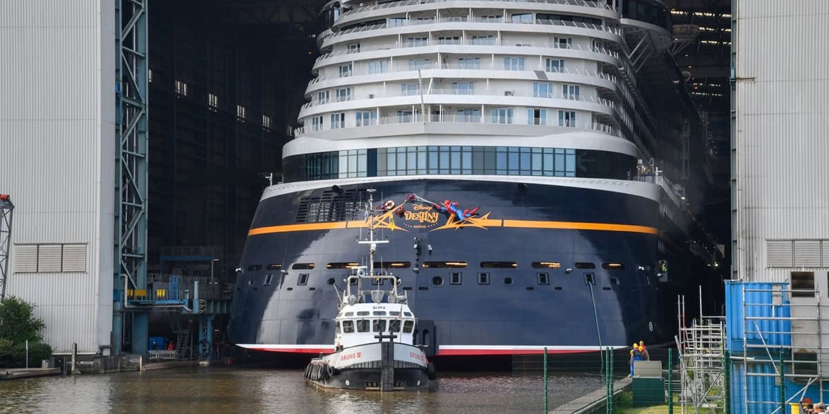 A large cruise ship, with "Disney" branding, is being moved out of a shipyard building. A small tugboat is positioned at the bow, helping guide the ship into the water. Workers and equipment are visible nearby.