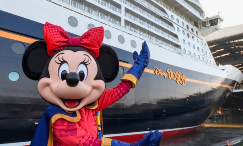 Minnie Mouse in a red and blue outfit stands in front of a large Disney cruise ship, smiling and waving, with the ship's name "Disney Wish" visible on the side as Disney trips get altered.