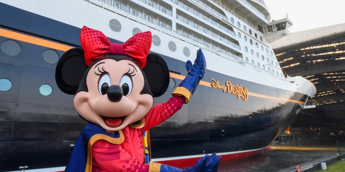 Minnie Mouse in a red and blue outfit stands in front of a large Disney cruise ship, smiling and waving, with the ship's name "Disney Wish" visible on the side as Disney trips get altered.
