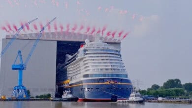 A large cruise ship exits a shipyard building, assisted by tugboats, with red fireworks and smoke in the sky above. Blue cranes and greenery are visible in the background near the water.
