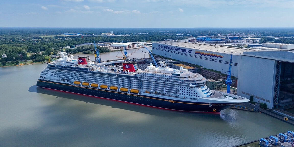 A large cruise ship docked at a shipyard near a white industrial building with the word "Papenburg" visible, surrounded by water and greenery under a partly cloudy sky as Disney trips take place.