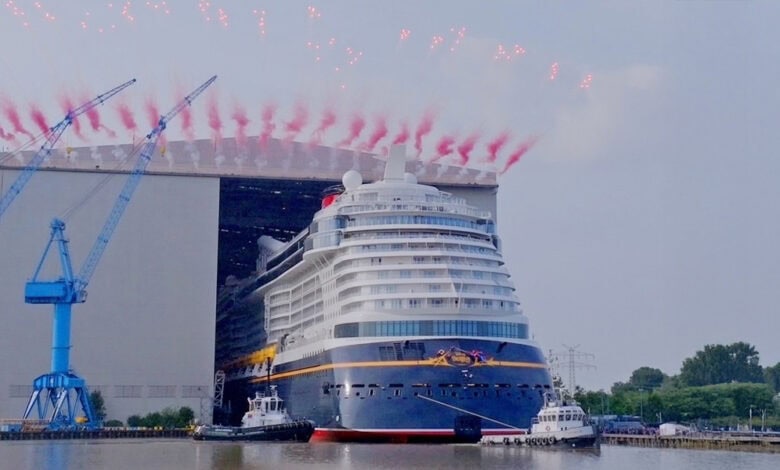 A large cruise ship exits a shipyard building, assisted by tugboats, with red fireworks and smoke in the sky above. Blue cranes and greenery are visible in the background near the water.