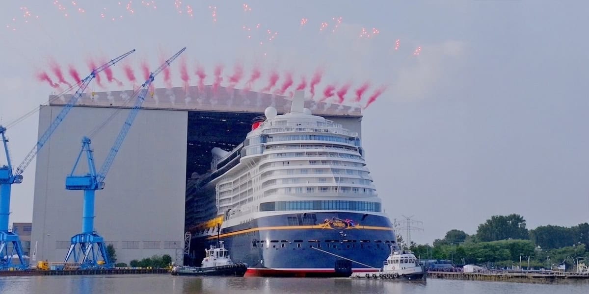 A large cruise ship exits a shipyard building, assisted by tugboats, with red fireworks and smoke in the sky above. Blue cranes and greenery are visible in the background near the water.