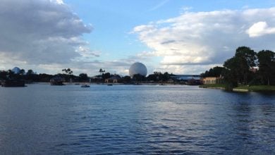 A large lake with calm water is in the foreground. Across the lake, trees and buildings line the shore, with Epcot's iconic white geodesic sphere visible in the distance under a partly cloudy blue sky at Disney World.