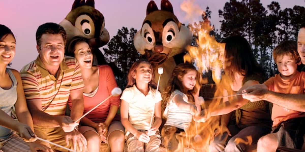 A group of people, including children, roast marshmallows over a campfire at sunset at Fort Wilderness, with two people dressed as chipmunk characters and trees in the background. Everyone appears to be smiling and enjoying the moment.