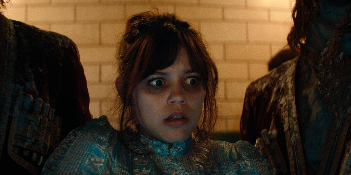 A young woman resembling Jenna Ortega, with dark hair and bangs, wears a high-collared, old-fashioned dress and looks ahead with a frightened expression. She is flanked by two shadowy figures in tattered clothing against a tiled background.