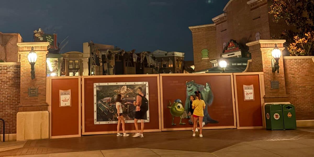 Two people stand near a construction wall featuring artwork of Sulley and Mike Wazowski from Monsters, Inc. at night. Another person stands nearby. Buildings and lamps are visible in the background.