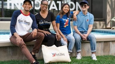 Four people sit by a fountain, smiling at the camera. They wear shirts with "Walt Disney Imagineering" logos, and a cream-colored bag with the same logo rests on the grass in front of them.