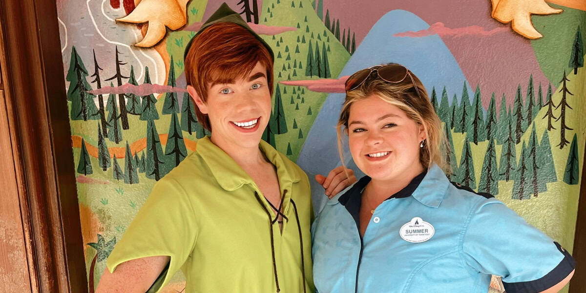 A person dressed as Peter Pan stands smiling next to a woman in a light blue uniform with a name tag, both posing in front of a colorful painted forest backdrop.