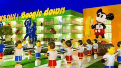 A vibrant outdoor scene at Pop Century Resort features large foosball player statues and a giant Mickey Mouse figure blowing a bubble, with colorful buildings and “Boogie Down” signage in the background.