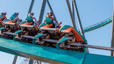 A group of people ride the green and orange Goliath Six Flags roller coaster as it climbs upward on the track against a clear blue sky. The riders appear excited and are securely seated in the coaster cars at Six Flags.