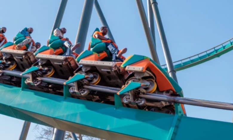 A group of people ride the green and orange Goliath Six Flags roller coaster as it climbs upward on the track against a clear blue sky. The riders appear excited and are securely seated in the coaster cars at Six Flags.