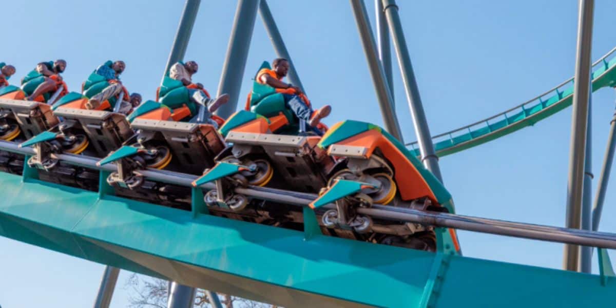 A group of people ride the green and orange Goliath Six Flags roller coaster as it climbs upward on the track against a clear blue sky. The riders appear excited and are securely seated in the coaster cars at Six Flags.