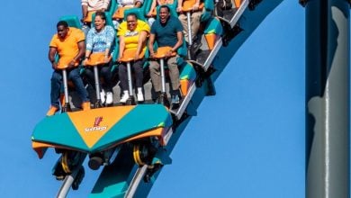 A group of people ride Goliath Six Flags, gripping the safety bars as they descend a steep track under a clear blue sky. The coaster car is orange and teal, with the Six Flags logo visible on the front.