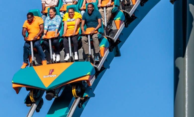 A group of people ride Goliath Six Flags, gripping the safety bars as they descend a steep track under a clear blue sky. The coaster car is orange and teal, with the Six Flags logo visible on the front.