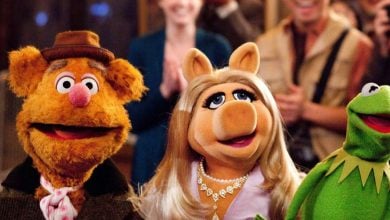 Fozzie Bear, Miss Piggy, and Kermit the Frog from The Muppets stand together smiling, with people clapping and cheering in the blurred background.
