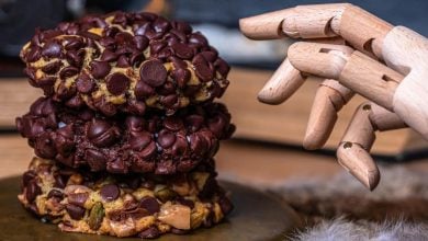 Three large cookies from Gideon's Bakehouse Disney Springs, covered in chocolate chips, are stacked on a surface next to a wooden articulated mannequin hand, with books and fur in the background.