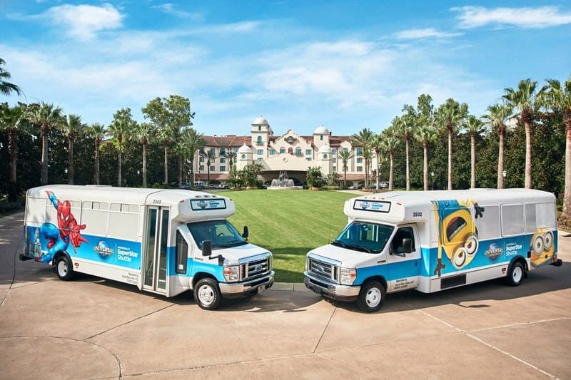Two white and blue shuttle buses with cartoon character graphics parked in front of a large hotel with palm trees and a green lawn under a blue sky.