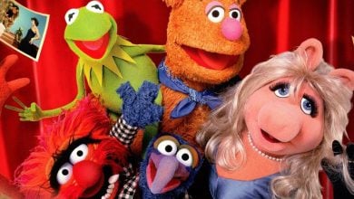Five colorful Muppet characters—including Kermit the Frog, Fozzie Bear, Miss Piggy, Animal, and Gonzo—pose happily together in front of a red curtain with photos pinned on the sides, celebrating The Muppets Disney Plus adventures.